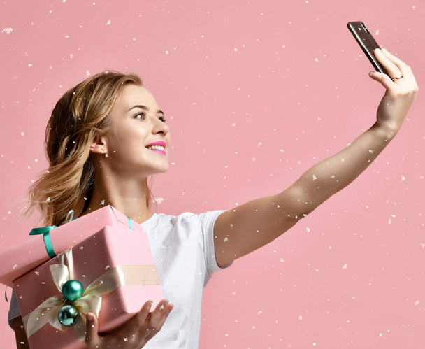 junge schöne Frau hält lächelnd Weihnachtsgeschenke in der Hand und macht Selfie-Foto mit ihrem Handy - Foto, Bild