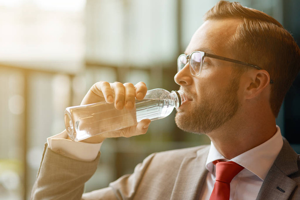Refrésquese. Primer plano de un joven empresario guapo sosteniendo una botella de agua y bebiéndola mientras mira a un lado
 - Foto, Imagen