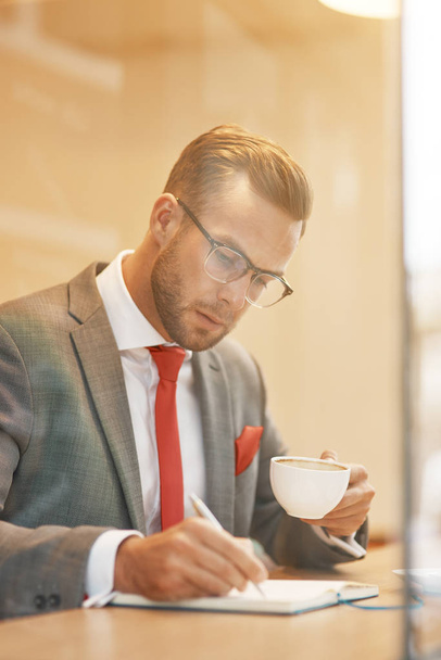 Мой ежедневный план. Талия молодого привлекательного бизнесмена, сидящего в кафе и пьющего кофе, планируя свой день
 - Фото, изображение