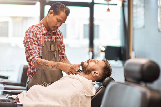 Professional movements. Masterful thai barber cutting a beard of a yougng handsome man while providing best service - Фото, изображение