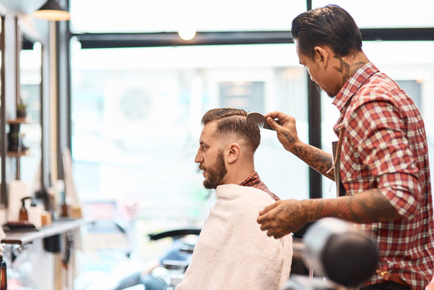 Looks good. Pleasant professional barber combing the hair of his male client while working in the barbershop - Foto, afbeelding