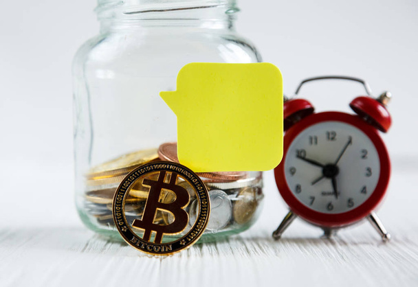 Bitcoins moneda de oro en el frasco de vidrio y despertador rojo en la mesa de madera blanca con etiqueta engomada de papel amarillo para el texto. Ahorro, concepto de inversión
 - Foto, Imagen