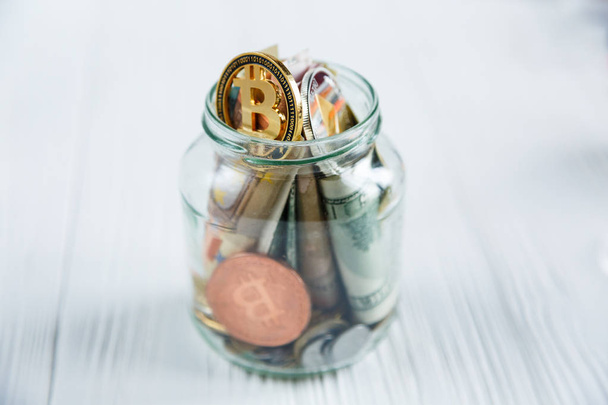 Bitcoins bronze golden coin in the glass jar on white wooden table. Set of cryptocurrencies with a real euro, dollars in the jar. Savings, investment, financial risk concept - 写真・画像