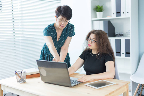 Business people, technology and communication concept - middle-aged woman showing something to another woman on laptop in office - Photo, Image