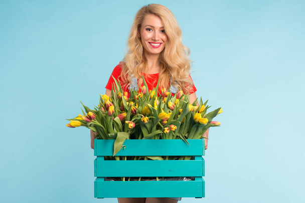 Beautiful blonde woman gardener holding box with tulips on blue background - Foto, imagen