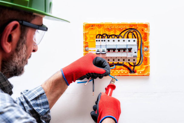 Technicien électricien avec casque, lunettes de protection et gants mains protégées, travaille avec la pince coupeur de fil dans un panneau électrique résidentiel
. - Photo, image