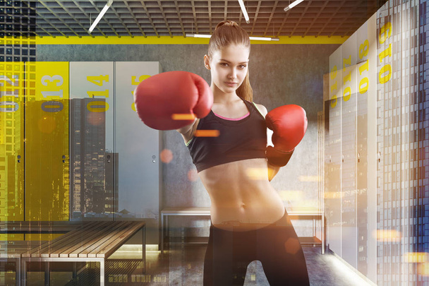 Woman with boxing gloves standing in locker room with concrete walls and floor, gray and yellow lockers and wooden benches. Toned image double exposure - Photo, image