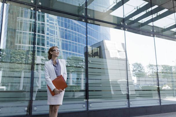 The young woman in gray office dress holds in hand folder with documents. Businesswoman standing next to the business center - Foto, afbeelding