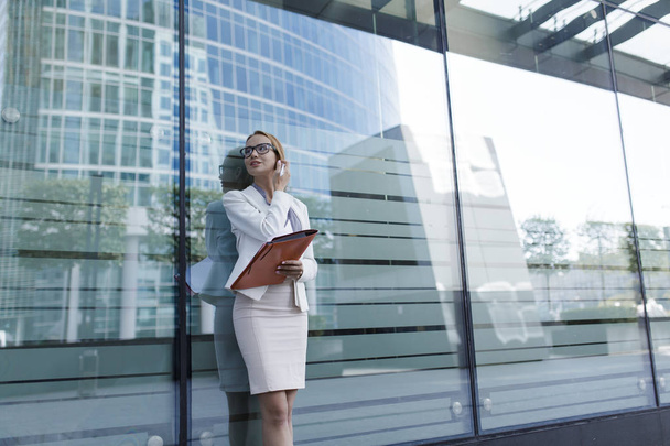 The young woman in gray office dress holds in hand folder with documents. Businesswoman standing next to the business center - Фото, изображение