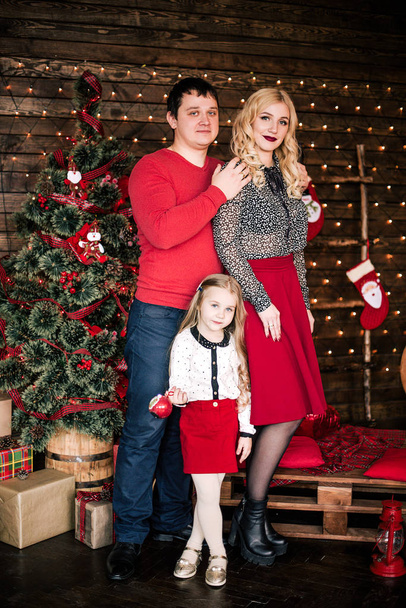 Молодая семья в красном весело проводит рождественские праздники, сидя на полу гостиной рядом с елкой, улыбаясь
 - Фото, изображение