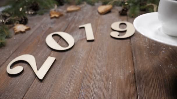 Frohe Weihnachten und ein gutes neues Jahr. 2019 Holzsymbole, Tannenzweige und Weihnachtsspielzeug auf Holzgrund. Frauenhände stellen Tasse Kaffee auf. Ansicht von oben - Filmmaterial, Video