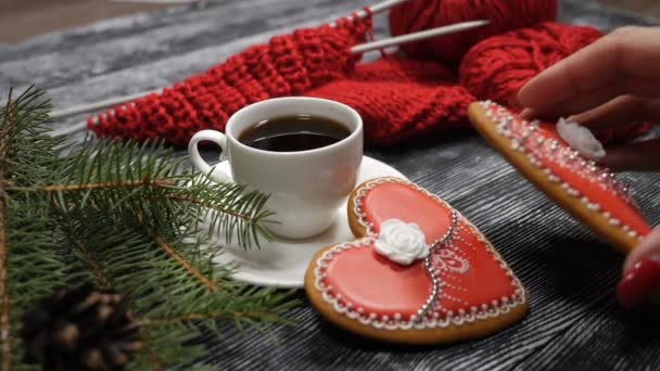 Cup of coffee and 2 red heartshaped ginger-snaps placed on a wooden background near fir tree branches and red yarn with knitting needle. Female hand puts one cookie on another. hd - Footage, Video