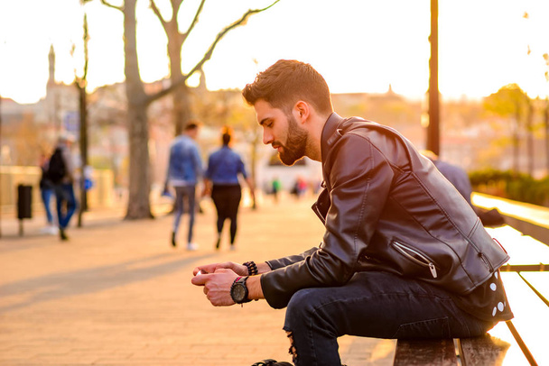 A handsome young hipster man sitting on a bench in the sunset and using his smartphone - 写真・画像