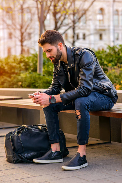 A handsome young hipster man sitting on a bench in the sunset and using his smartphone - Фото, изображение