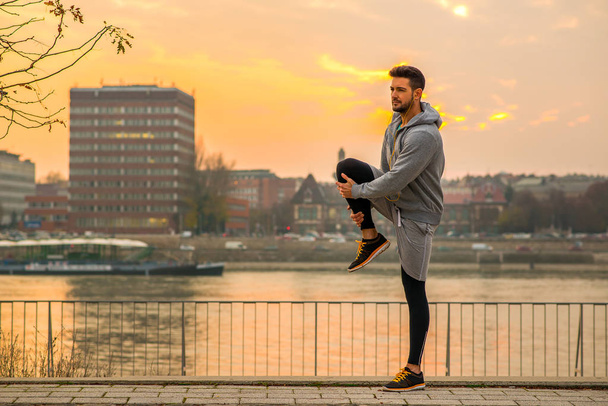 A handsome young man stretching and warming up on the riverside with the sun setting behind him - Foto, imagen