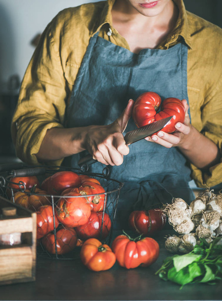 Woman in linen apron cutting ripe heirloom tomato and cooking tomato sauce, canned tomatoes or pasta with basil and garlic at kitchen counter. Healthy cooking, slow food or comfort food - Photo, image