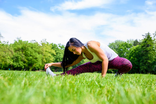 A beautiful young sporty girl stretching on a field in a park while wearing a leggings and a top - Valokuva, kuva