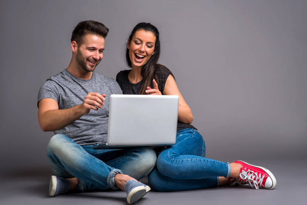 A beautiful young couple sitting on the ground and looking at a laptop and smiling - Фото, изображение