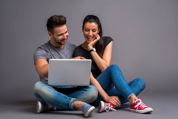 A beautiful young couple sitting on the ground and looking at a laptop and smiling - Foto, Imagem