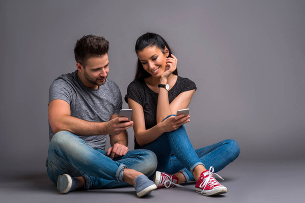 A beautiful young couple sitting on the ground and showing their smartphones to each other  - Foto, Imagen