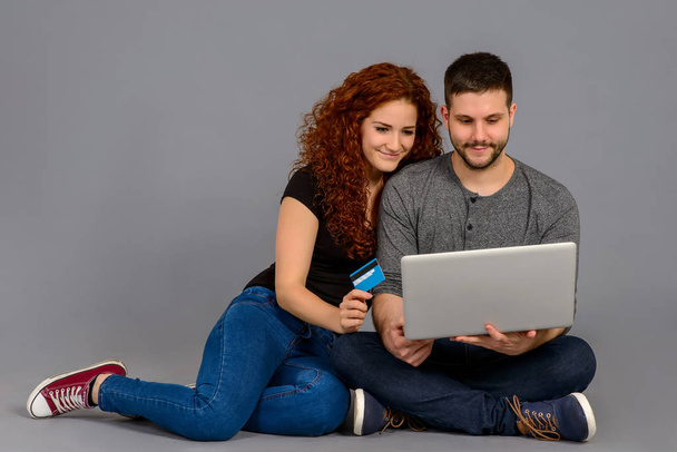 A handsome young guy holding a laptop while his girlfriend showing a credit card and they both smiling and sitting on the ground - Fotografie, Obrázek