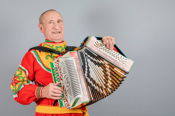 Portrait of a man in a national costume, grandfather plays the accordion. On a gray background - Photo, Image