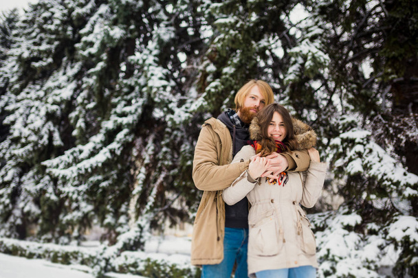 Young Caucasian boy and girl in love heterosexual couple have a date outdoors in a winter park on the background of a snowy conifer standing in an embrace. Man hugging woman valentines day - Photo, Image