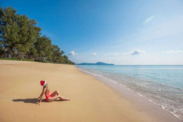 Young woman with long hair in red swimsuit and santa claus hat sitting on the wide tropical beach without people near the sea with beautiful blue water and sky on Phuket island,Thailand - 写真・画像