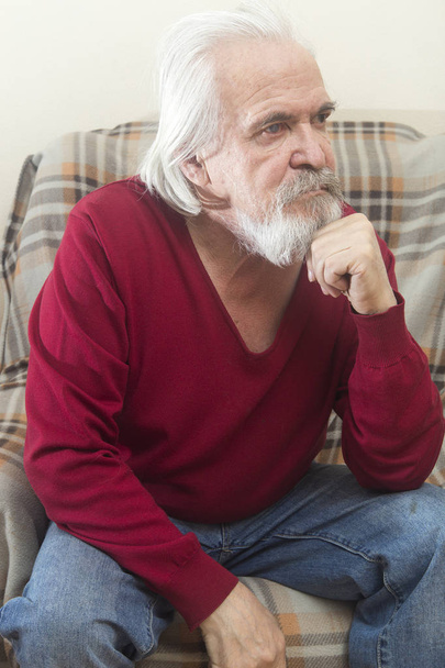 A handsome sick old man with a beard and a mustache with long white hair in a nursing home sits in an armchair all alone and reflects on the past years - Foto, Imagem
