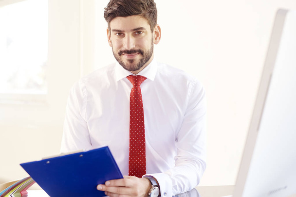 Portrait of smiling young businessman holding clipboard in his hand while sitting at office desk and looking at camera. - Foto, imagen