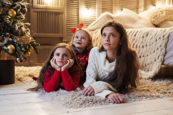 Three cute young sisters lying on the floor between Christmas tree and bed, having fun, smiling. Girls wearing red and white sweaters. - Fotoğraf, Görsel
