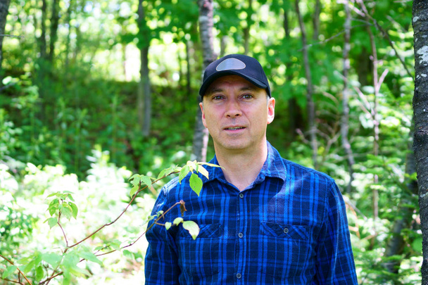 A caucasian curious man is looking away while hiking in the forest wearing a blue check shirt and a black hat.                                - Photo, Image