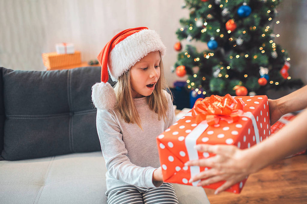 Amazed child holds present and looks at it. She keeps mouth opened. Girl wears Christmas hat. Adult supports her by holding present with hands. They are in decorated room. - Foto, afbeelding