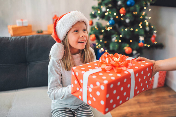 Beautiful little girl holds present with both hands and looks up. She is excited and happy. Adult holds present as well. There are Christmas tree and sofa on background. - Foto, Bild