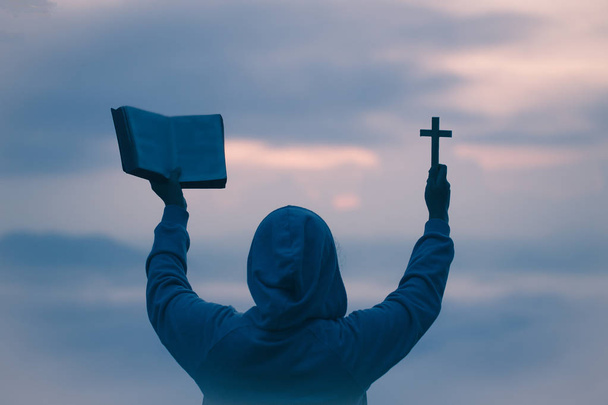 Sylwetka kobiety ręce trzymając drewniany krzyż i Biblii na wschód tło, krucyfiks, Symbol wiary. - Zdjęcie, obraz