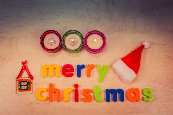 Merry Christmas concept met tekst op achtergrond, brandende kaarsen, huisje van glas en kerstman hoed - Foto, afbeelding