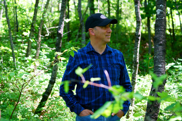 Valkoihoinen mies katsoo poispäin vaeltaessaan metsässä yllään sininen shekkipaita ja musta hattu.
. - Valokuva, kuva
