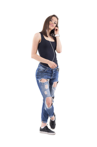 Young trendy woman in black top and torn jeans with earphones posing and looking at camera. Full body isolated on white background.  - Photo, Image