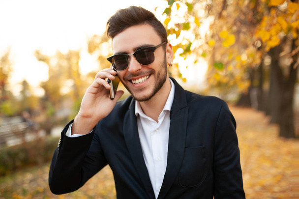 Handsome young stylish male businessman happy and smiling talking on the phone on a walk in the autumn park on the background of yellow leaves - Фото, изображение