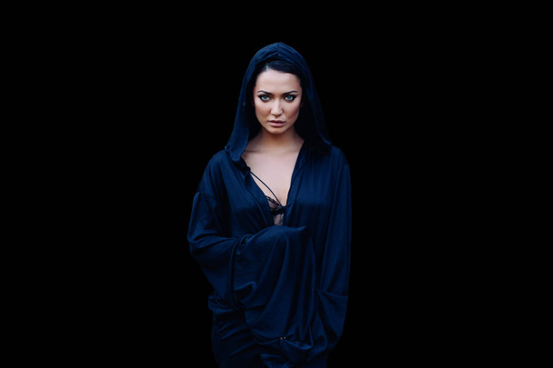 Young beautiful woman with a black hair and in the dark blue cloak with hood at the black background - Zdjęcie, obraz