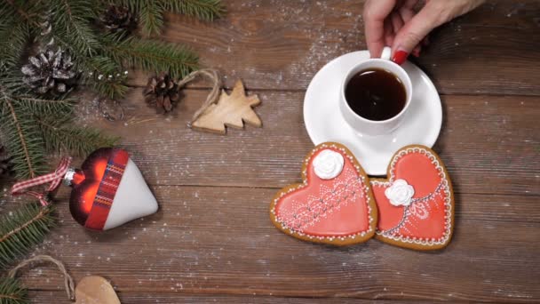 Καλά Χριστούγεννα και Ευτυχισμένο το νέο έτος έννοια. Καρδιά σχήμα cookies, κλαδιά δέντρων ελάτης και Χριστούγεννα παιχνίδια στο ξύλινο υπόβαθρο. Γυναικείο χέρι παίρνει μακριά ένα φλιτζάνι καφέ. Βίντεο καρτ ποστάλ. HD - Πλάνα, βίντεο