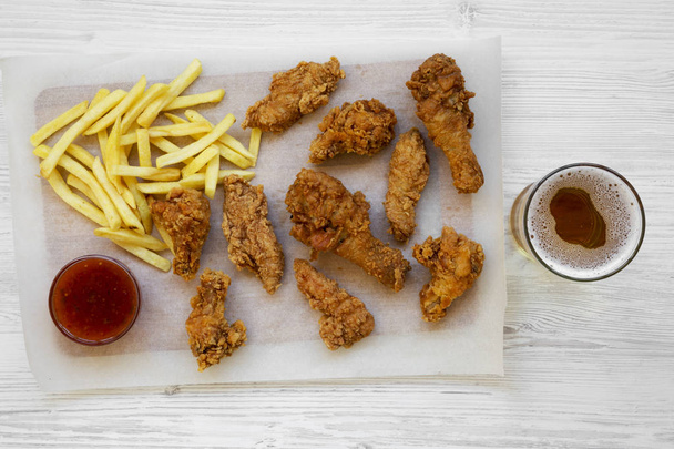 Fried chicken drumsticks, spicy wings, French fries and chicken strips with sour-sweet sauce and cold beer over white wooden background, top view. Flat lay, overhead, from above. - 写真・画像