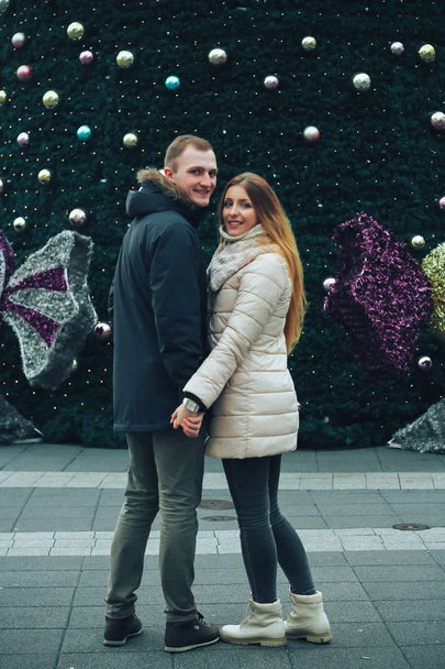 寒い冬の夜に楽しむ美しい若いカップル。彼らは通りに立ってクリスマスツリーの装飾品を見ています。背景にカラフルな街路灯。新年のテーマ. - 写真・画像