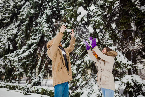 Young Caucasian boy with a beard and a girl have a date outdoors in the winter park against the background of a snowy conifer tree play snowballs, throw snow, hang themselves and play winter games. - Photo, Image
