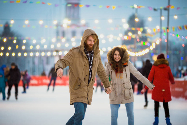 Young couple in love Caucasian man with blond hair with long hair and beard and beautiful woman have fun, active date ice skating on the ice arena in the evening city square in winter on Christmas Eve - Photo, Image