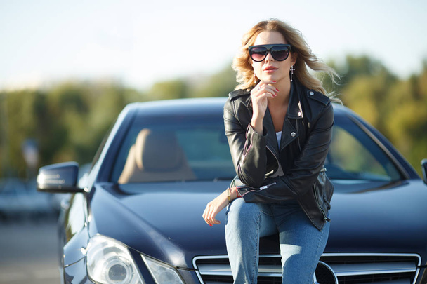Photo of young woman in sunglasses sitting on hood of black car - Foto, afbeelding