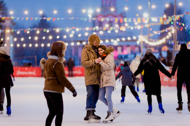若いカップルが長い髪の金髪の白人男性を愛し、ひげと美しい女性がクリスマスイブに冬の夜市広場でアイス アリーナでスケートを楽しみ、アクティブな日付氷を持って - 写真・画像