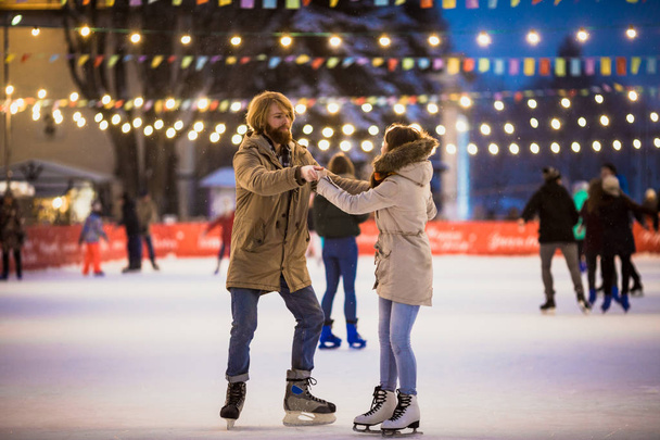 Young couple in love Caucasian man with blond hair with long hair and beard and beautiful woman have fun, active date ice skating on the ice arena in the evening city square in winter on Christmas Eve - Foto, afbeelding