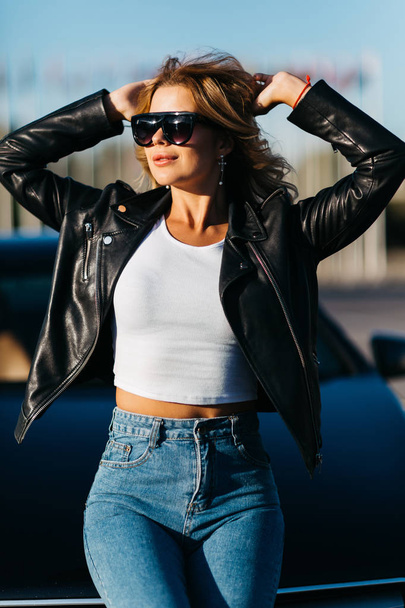 Portrait of young blonde girl in sunglasses standing next to black car - Foto, Imagen