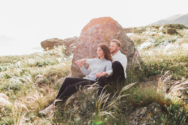 Happy young loving couple sitting in feather grass meadow, laughing and hugging, casual style sweater and jeans - Photo, Image